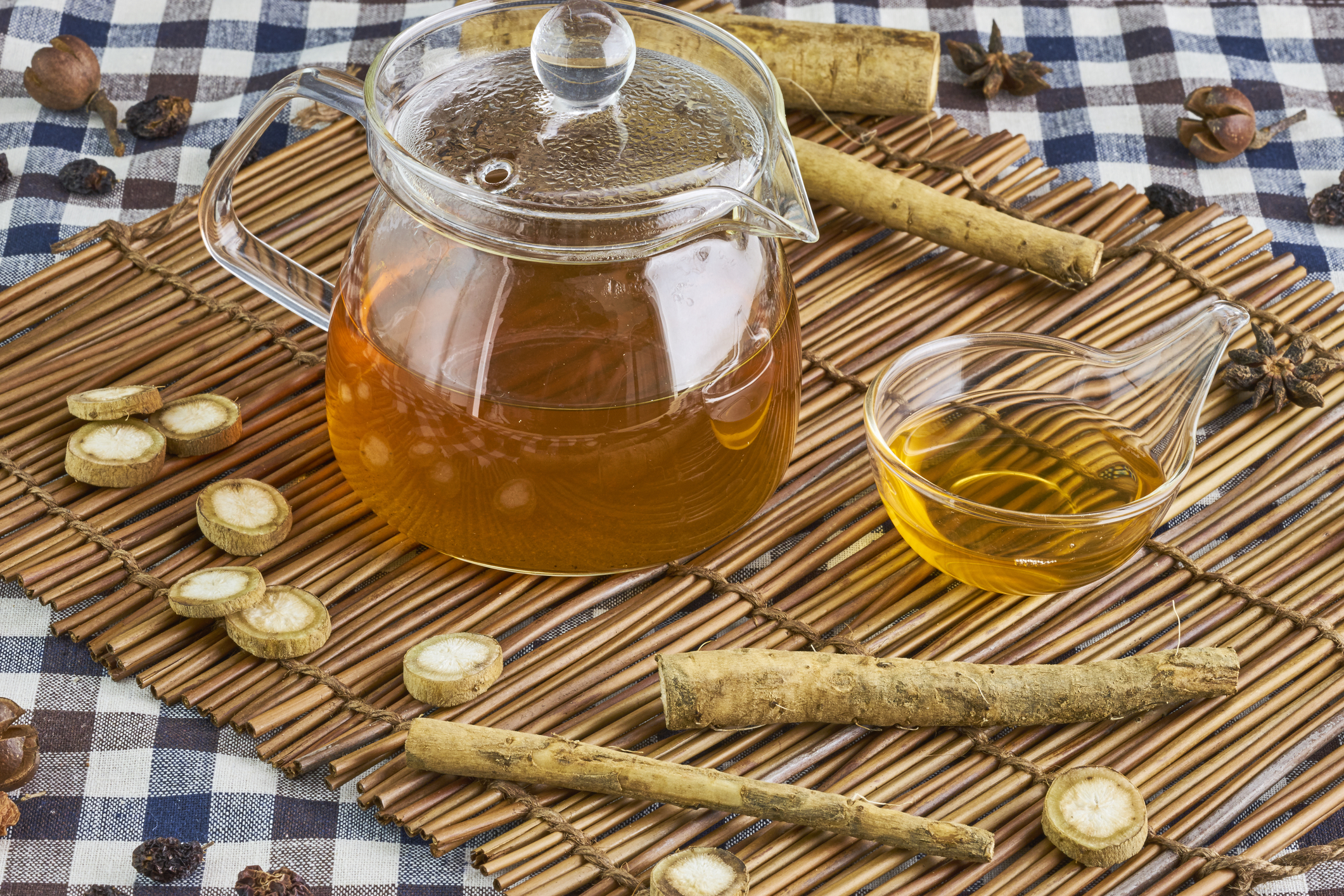 Small glass teapot of golden tea surrounded by burdock roots on a bamboo mat.