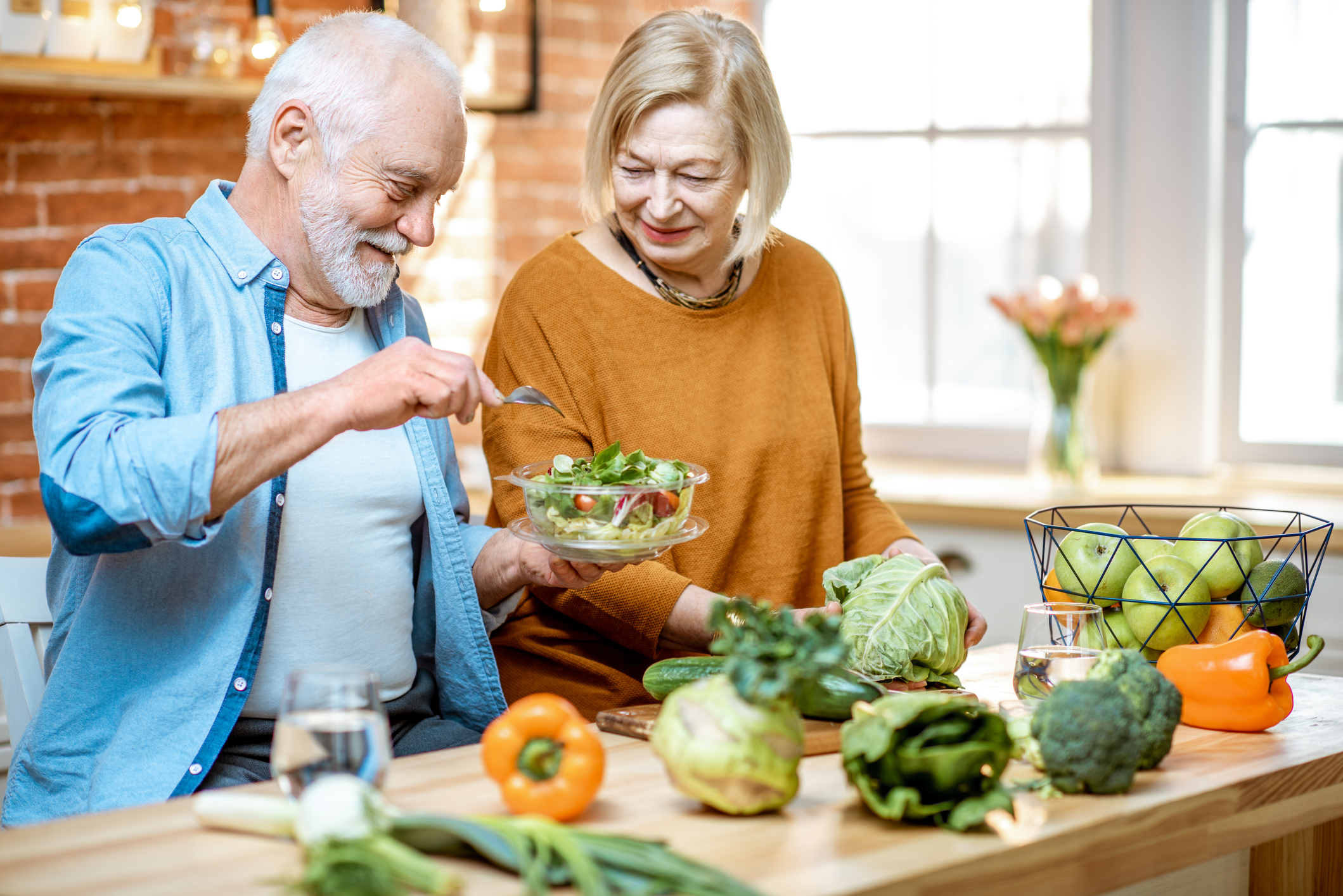 A senior man eating from a salad while a senior woman looks on.