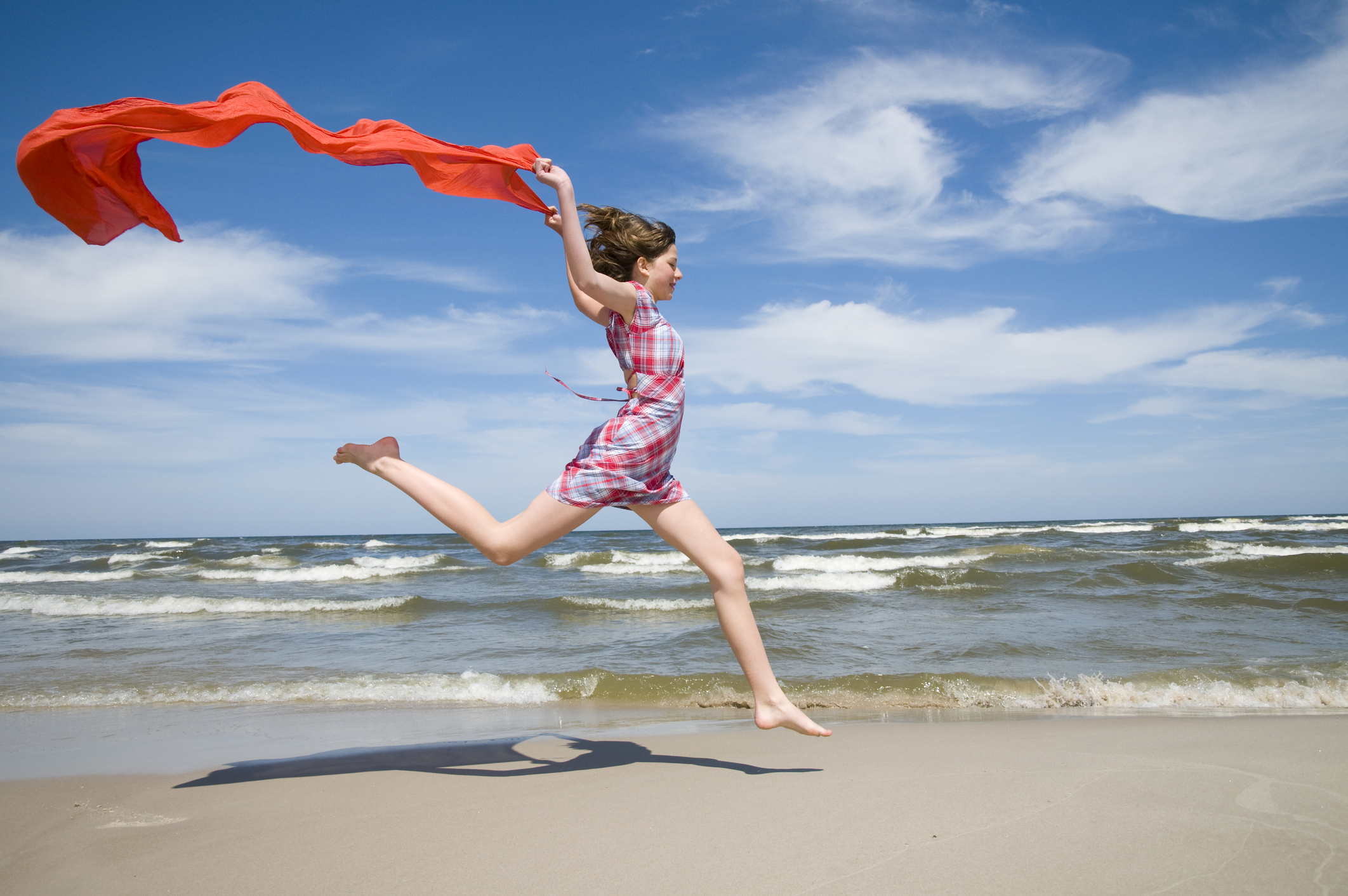 A teenage girl runs along a beach trailing a red cloth behind her.