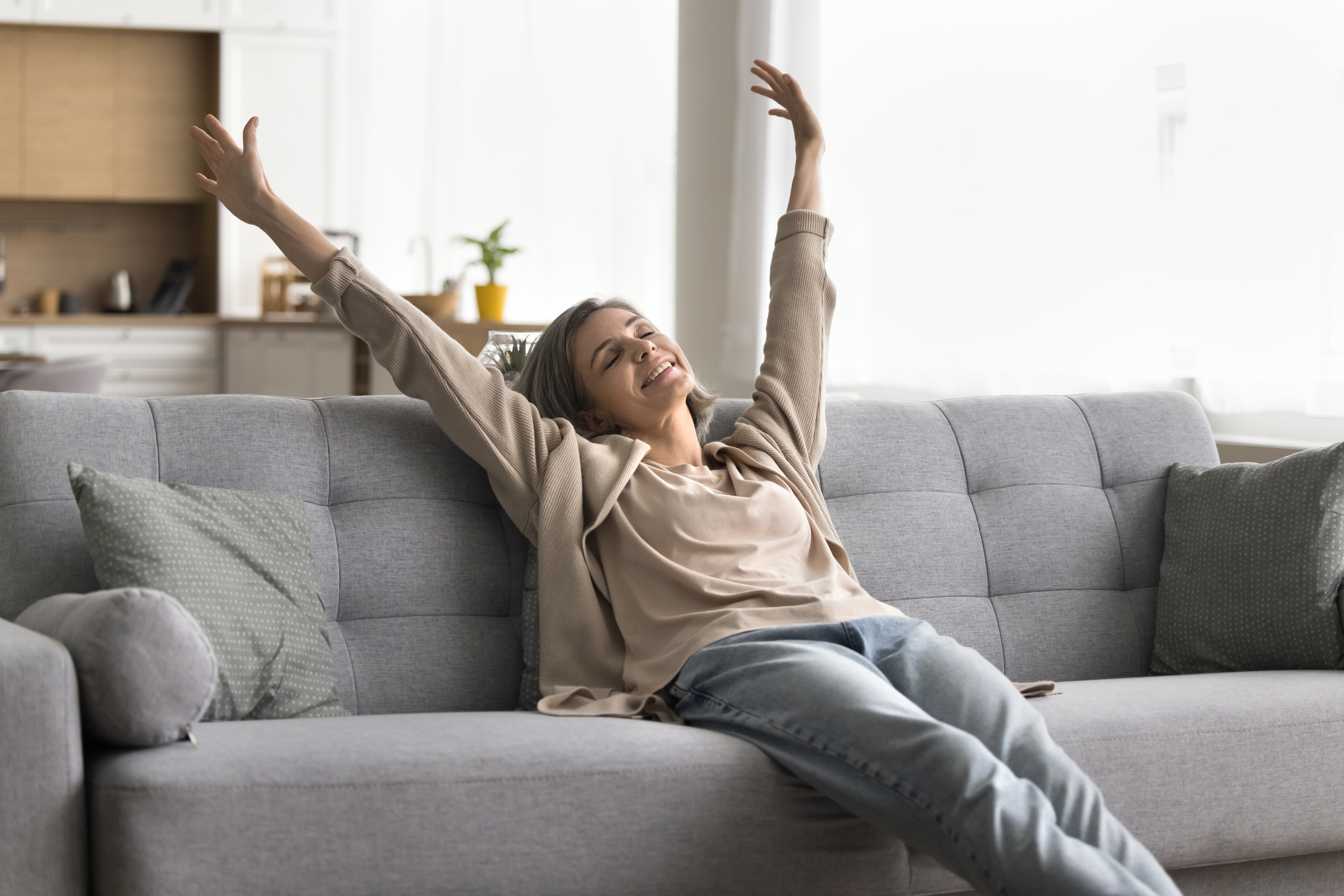 Mid-life woman sitting on a grey couch stretching her arms overhead.