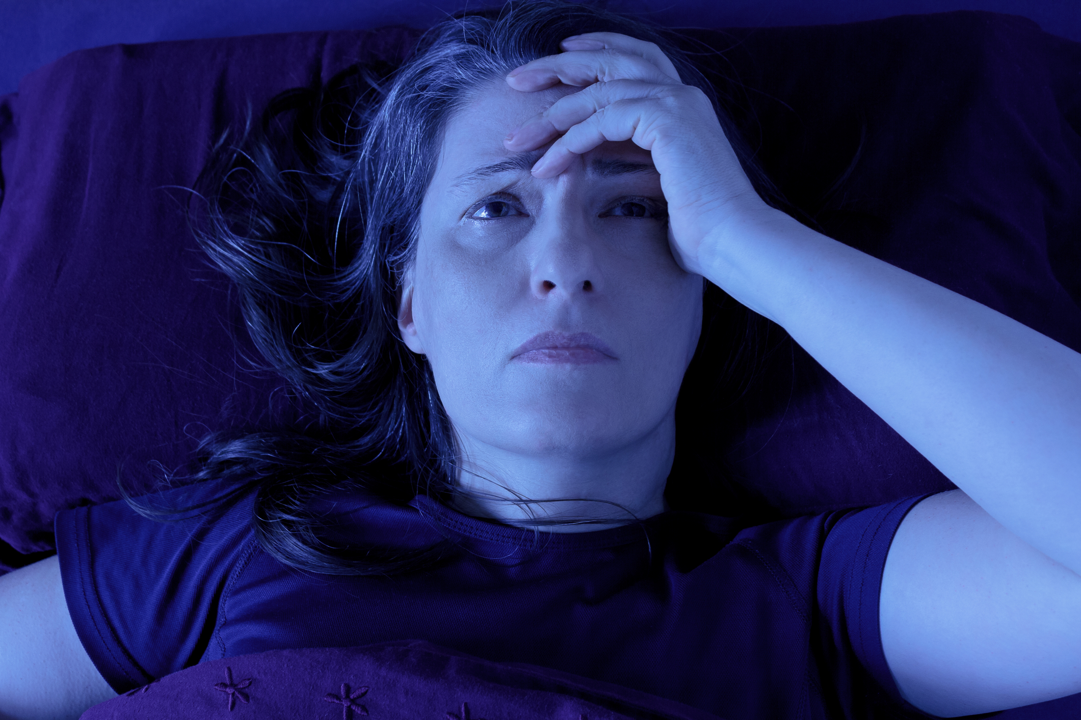A worried-looking woman lying in bed at night, holding her head in her left hand. 
