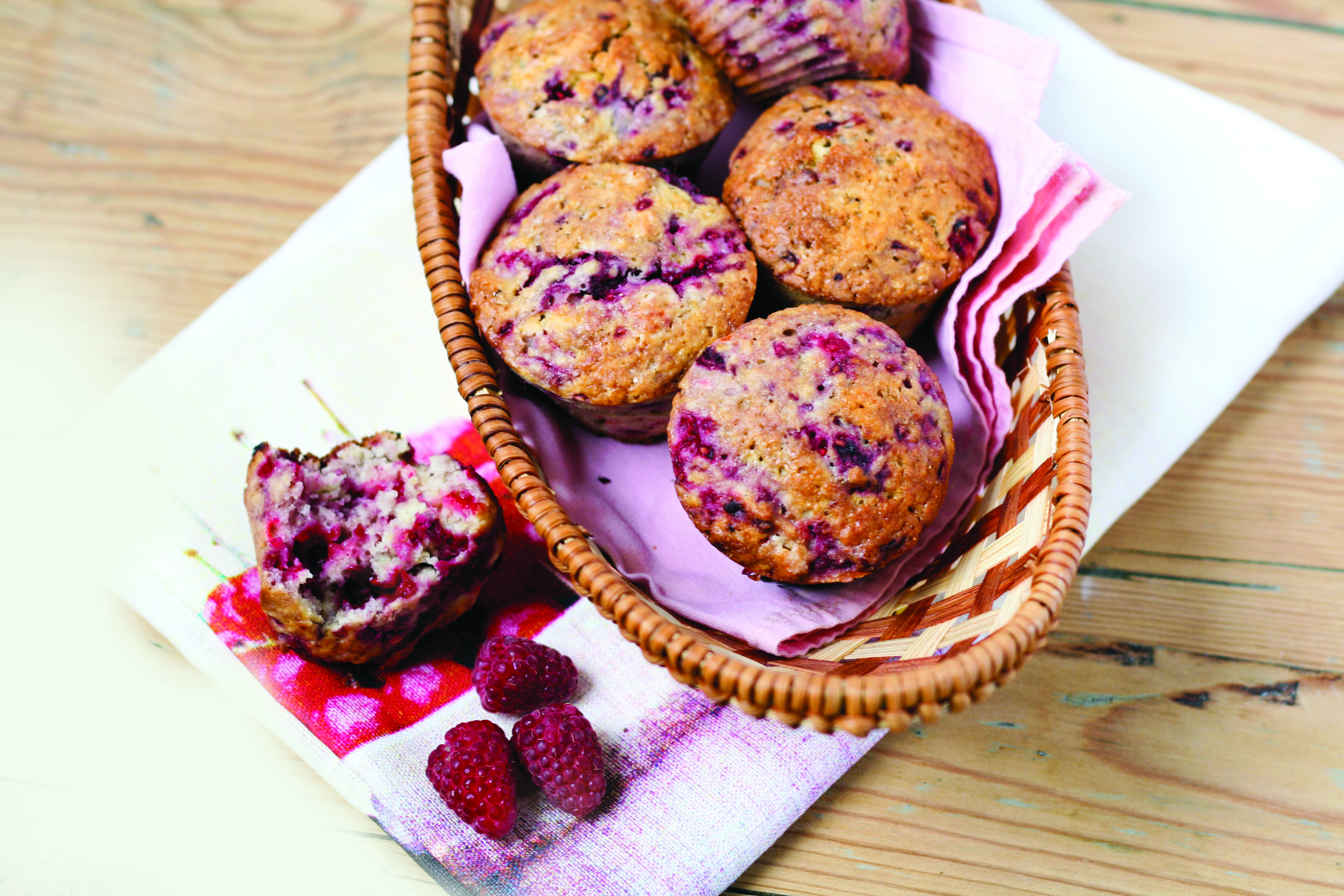Raspberry muffins in a basket, placed on a white cloth alongside a broken muffin and raspberries.