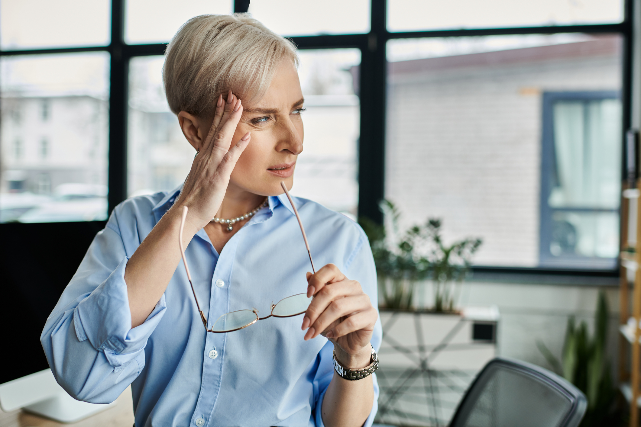 A caucasian middle-aged woman with short hair calmly holds her glasses while touching her temple.
