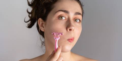 A young woman holds a razor to her cheek to remove excess hair growth caused by PCOS.