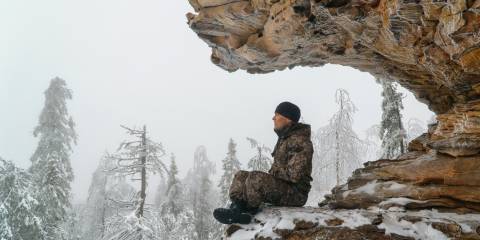 Man dressed in camouflage and a hat meditating on a mountain in winter. 