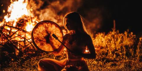a shamanic woman beating a drum by the fire