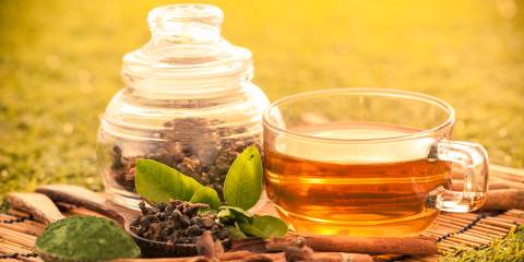 Brewed green tea in a glass cup on a bamboo placemat with dried green tea in a jar next to it outdoors on the grass.