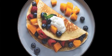 A pancake wrapped around cantaloupe chunks, raspberries, and blueberries, with a dollop of yogurt on top.