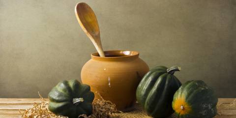 Whole acorn squash on a rustic table next to an earthen vase with a wooden spoon i it.