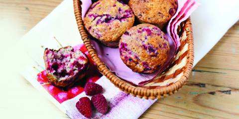Raspberry muffins in a basket, placed on a white cloth alongside a broken muffin and raspberries.