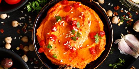 Top view of Red Hummus in a dark colored bowl surrounded by ingredients and foods to dip. Dark background.