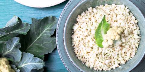 Cauliflower rice in a blue bowl on a blue table next to a head of cauliflower with leaves.