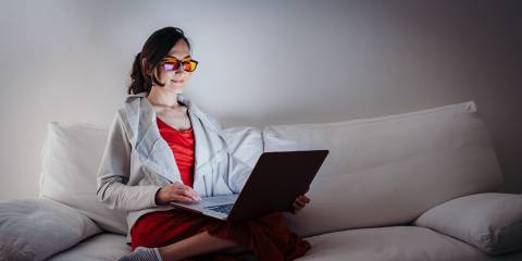 Young woman with blue light blocking glasses yellow lenses working with laptop on white sofa