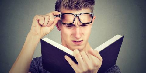 A young man lifting his glasses straining to read a book.