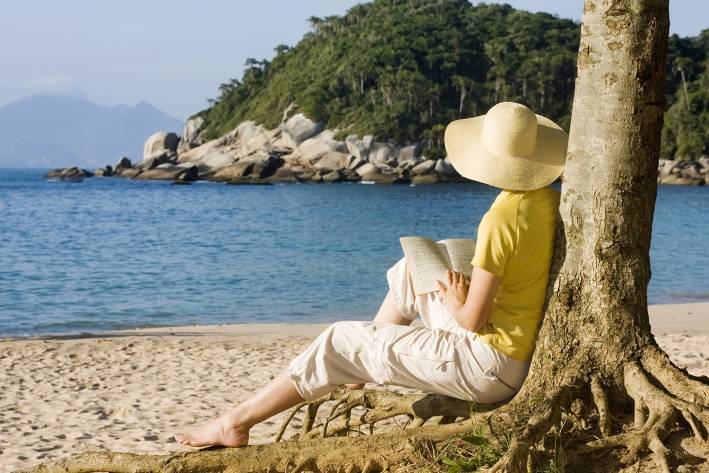 a woman reading, sitting against a tree on the beach