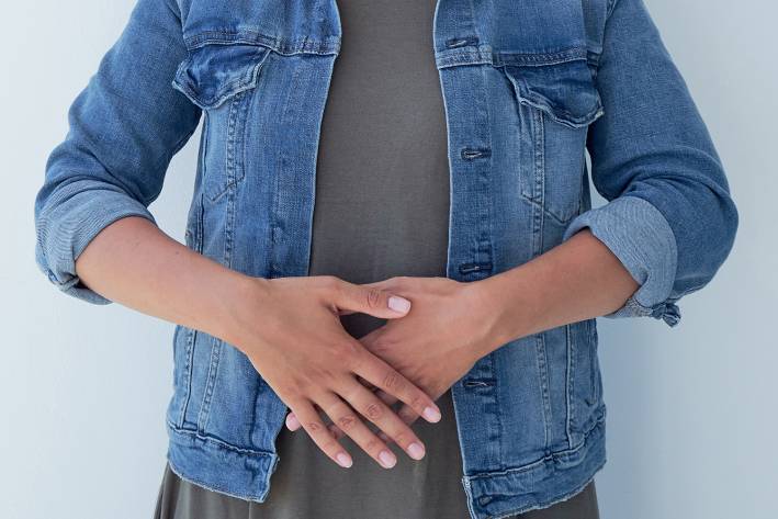 A woman in a gray shirt and jean jacket with hands crossed over her abdomen.