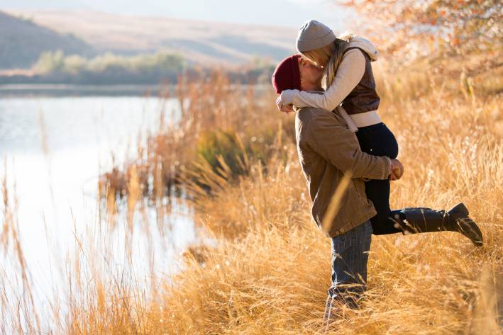 Man holding a woman in the air beside a mountain lake in autumn.