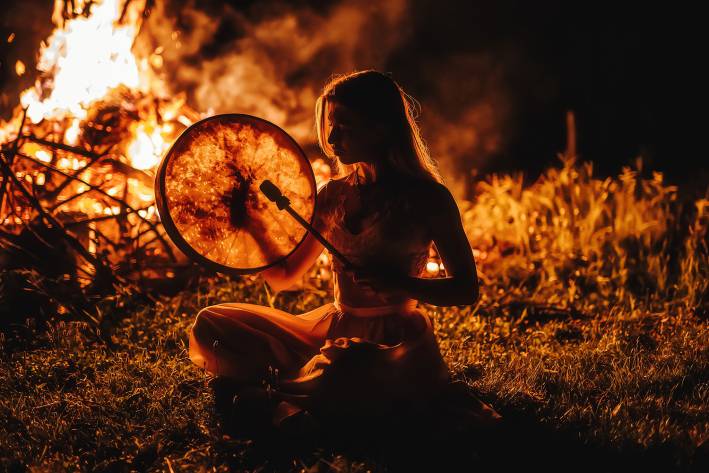 a shamanic woman beating a drum by the fire