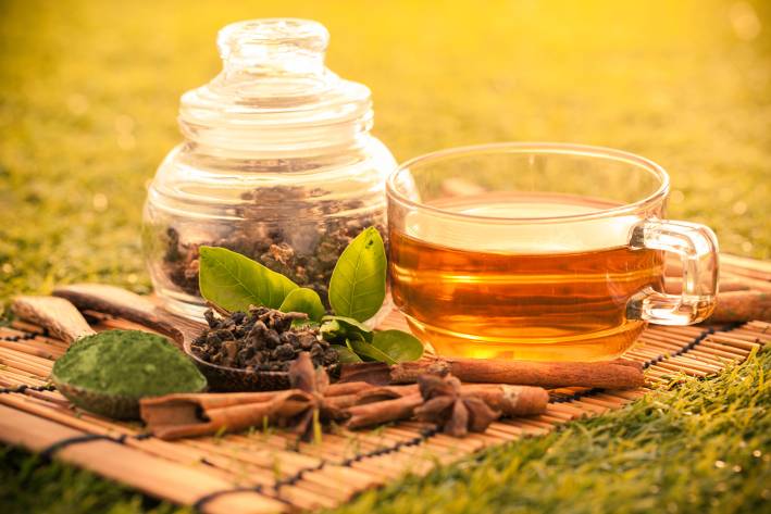 Brewed green tea in a glass cup on a bamboo placemat with dried green tea in a jar next to it outdoors on the grass.