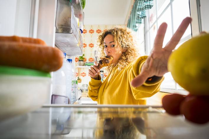 a woman eating while grabbing food out of the fridge