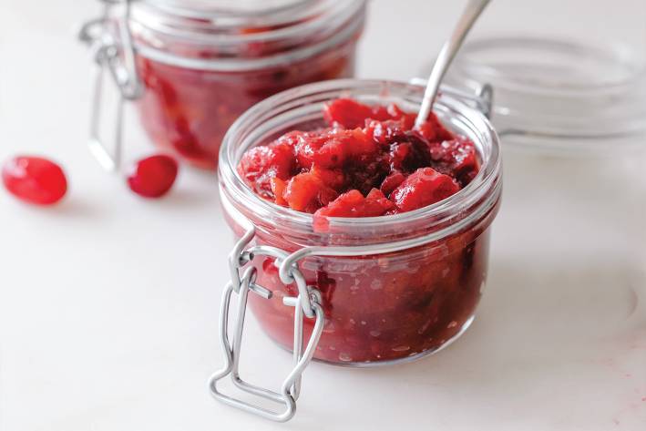 A small flip top glass jar of cranberry chutney with a small serving spoon in it, white background.