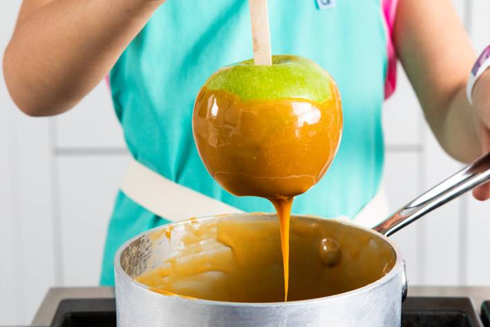 A woman dipping a Granny Smith apple in a pot of caramel to make caramel apples.