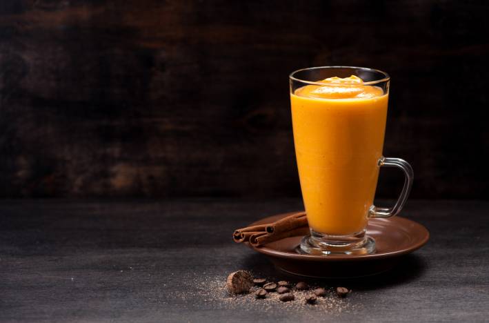 A glass cup filled with pumpkin spice smoothie next to cinnamon sticks. 