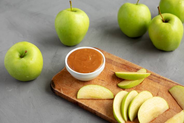 Caramel dip in a bowl on a cutting board with sliced green apples. 