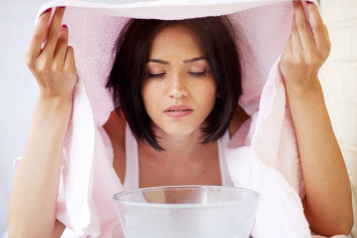 A woman relaxing during a facial steam treatment over a bowl with towel over her head.