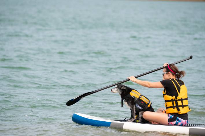 A woman with a yellow life vest is seated and paddling a paddleboat while an older black dog with a white muzzle, wearing a matching yellow safety vest, sits in front of her.