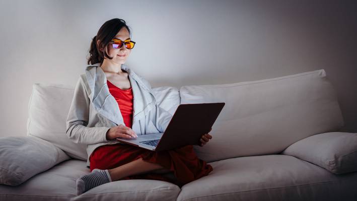 Young woman with blue light blocking glasses yellow lenses working with laptop on white sofa