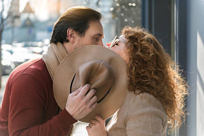 Romantic mature couple standing outdoors hiding behind a hat and kissing.