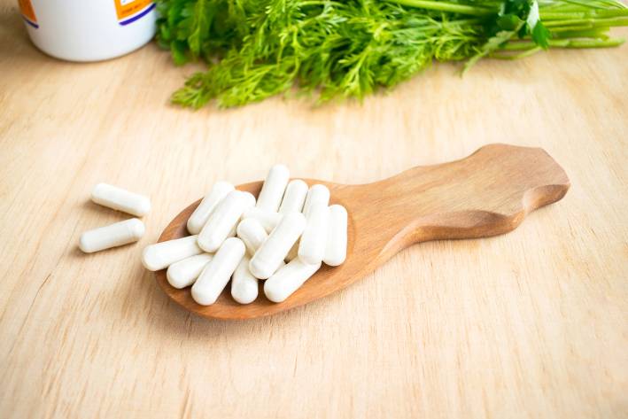 Vitamins, supplements in spoon on wooden table with green herbs in the background.