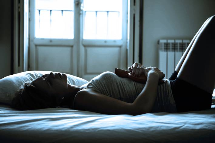 A woman laying down in the dark with a hot water bottle on her abdomen