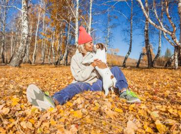 Woman with a winter hat on sitting in yellow leaves holding her little white and brown dog.