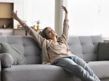 Mid-life woman sitting on a grey couch stretching her arms overhead.