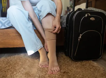 Woman sitting on bed next to a suitcase, pulling up compression sock.