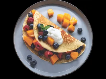 A pancake wrapped around cantaloupe chunks, raspberries, and blueberries, with a dollop of yogurt on top.