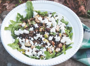 A white bowl of salad made up of arugula, goat cheese and other ingredients.