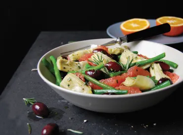 Green Bean, Artichoke, & Grapefruit Salad with Olives and Rosemary in a white bowl with a cut orange in the background. Black background.