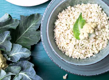 Cauliflower rice in a blue bowl on a blue table next to a head of cauliflower with leaves.