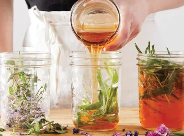 A woman in a white apron pouring honey into jars of herbs.