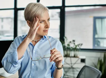 A caucasian middle-aged woman with short hair calmly holds her glasses while touching her temple.